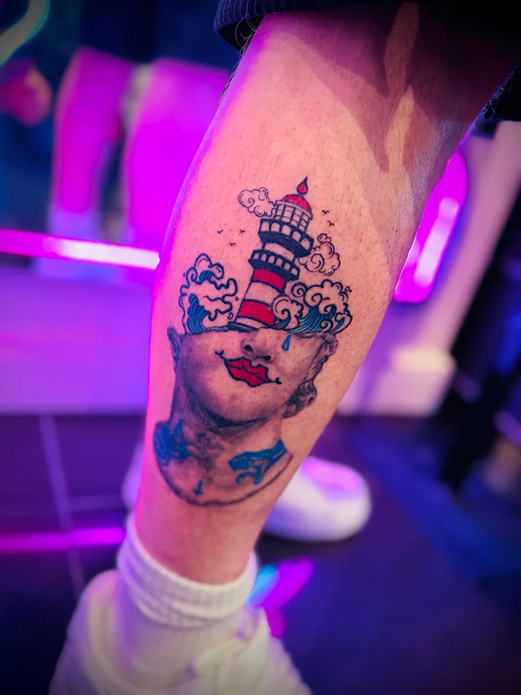 A photograph of a mashup tattoo combining a black and grey statue head with a full colour traditional style lighthouse